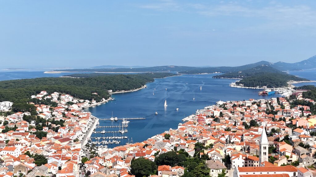 Drone view of Mali Lošinj Bay from the hill near the church – a well-sheltered North Adriatic sailing destination with turquoise waters, a vibrant marina, a scenic promenade, and the famous Museum of Apoxyomenos reacghable with charter from Pula or Krk island